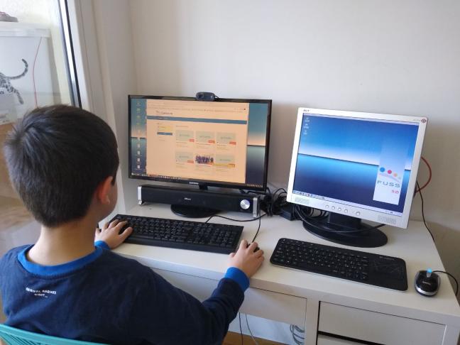 A school student sitting at a desk at home, with two computer screens showing the FUSS open source software 